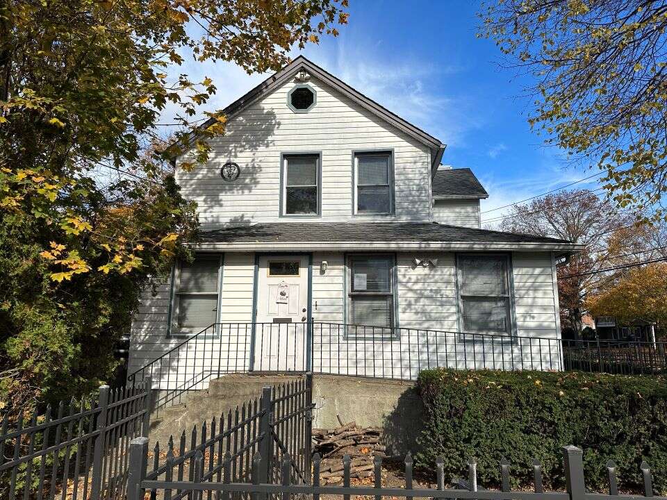 A two-story white house with a gabled roof, surrounded by trees and a black metal fence.