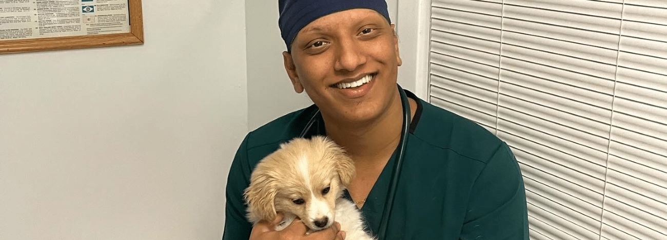 Person in scrubs smiling and holding a small, fluffy puppy indoors.